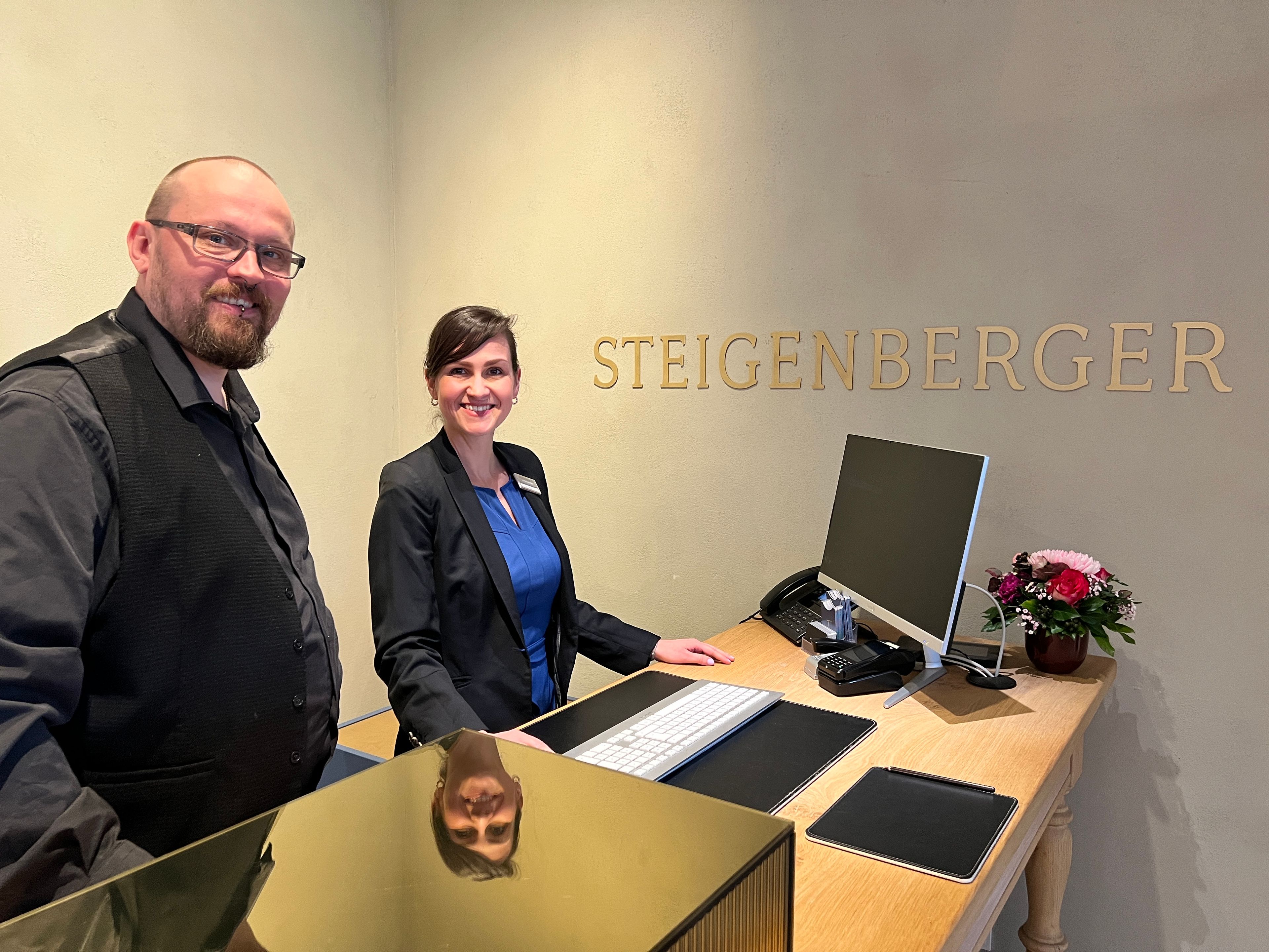 Zwei freundliche Mitarbeitende am Empfangsbereich eines Hotels oder Unternehmens mit moderner Rezeption und persönlichem Kundenservice.